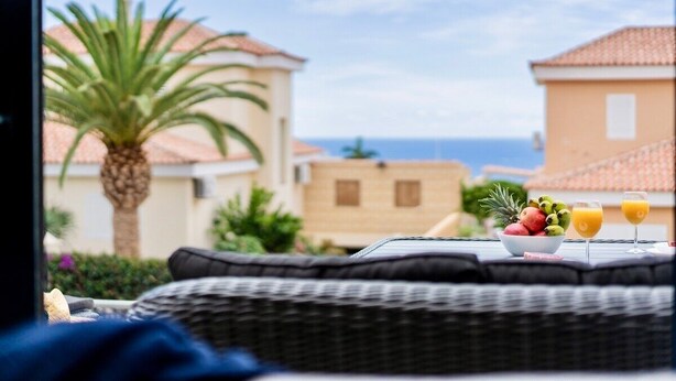 A wicker sofa with a view of a coastal town, a bowl of fruit, and two glasses of orange juice on a table. - Charming Villa Mirador del Duque (Costa Adeje)