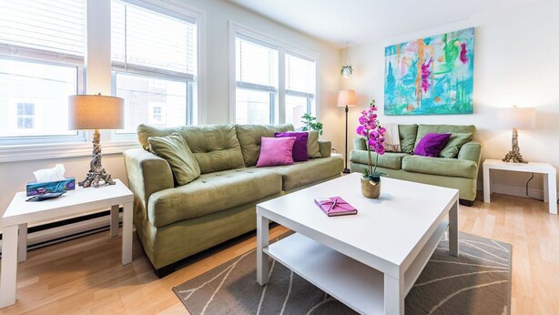 A modern living room with two green sofas, a white coffee table, and a colorful abstract painting on the wall. - Jellybean Dream upstairs apt: driveway, 4 bedrooms & near Bannerman Park! (St. John's)