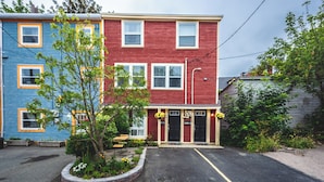 A two-story building with a red and blue exterior, a parking lot, and a small garden area with a tree and flowers. - Jellybean Dream upstairs apt: driveway, 4 bedrooms & near Bannerman Park! (St. John's)