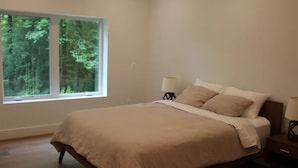 A bedroom with a large window, a bed, two bedside tables with lamps, and a view of trees outside. - The Wooded Retreat (McLean)