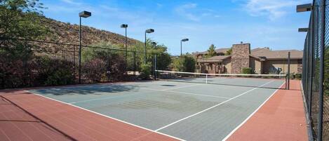 An outdoor tennis court with a net, surrounded by a fence and street lights.