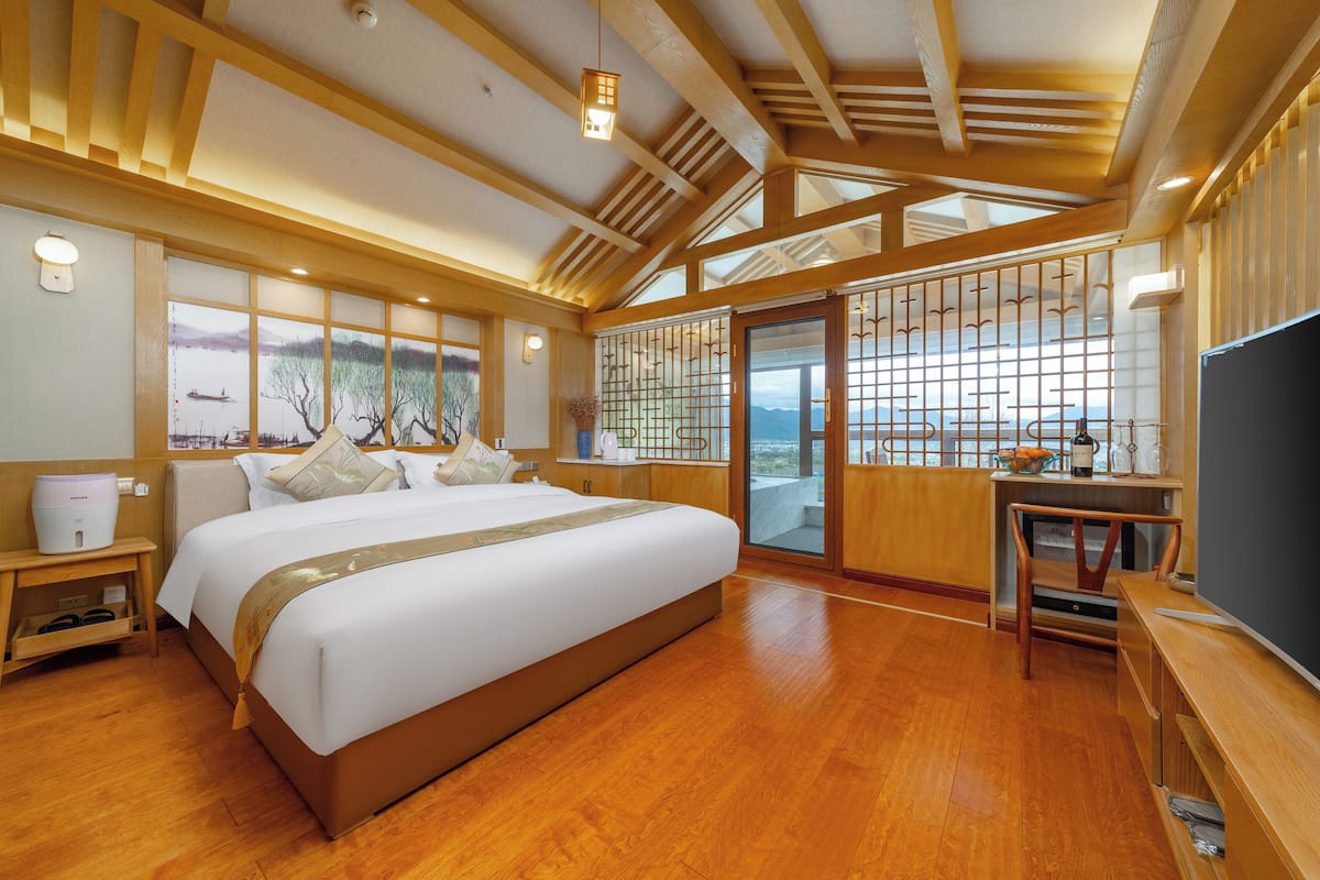 A spacious bedroom with a large bed, wooden flooring, a view of the outdoors, and a sliding door leading to a balcony.