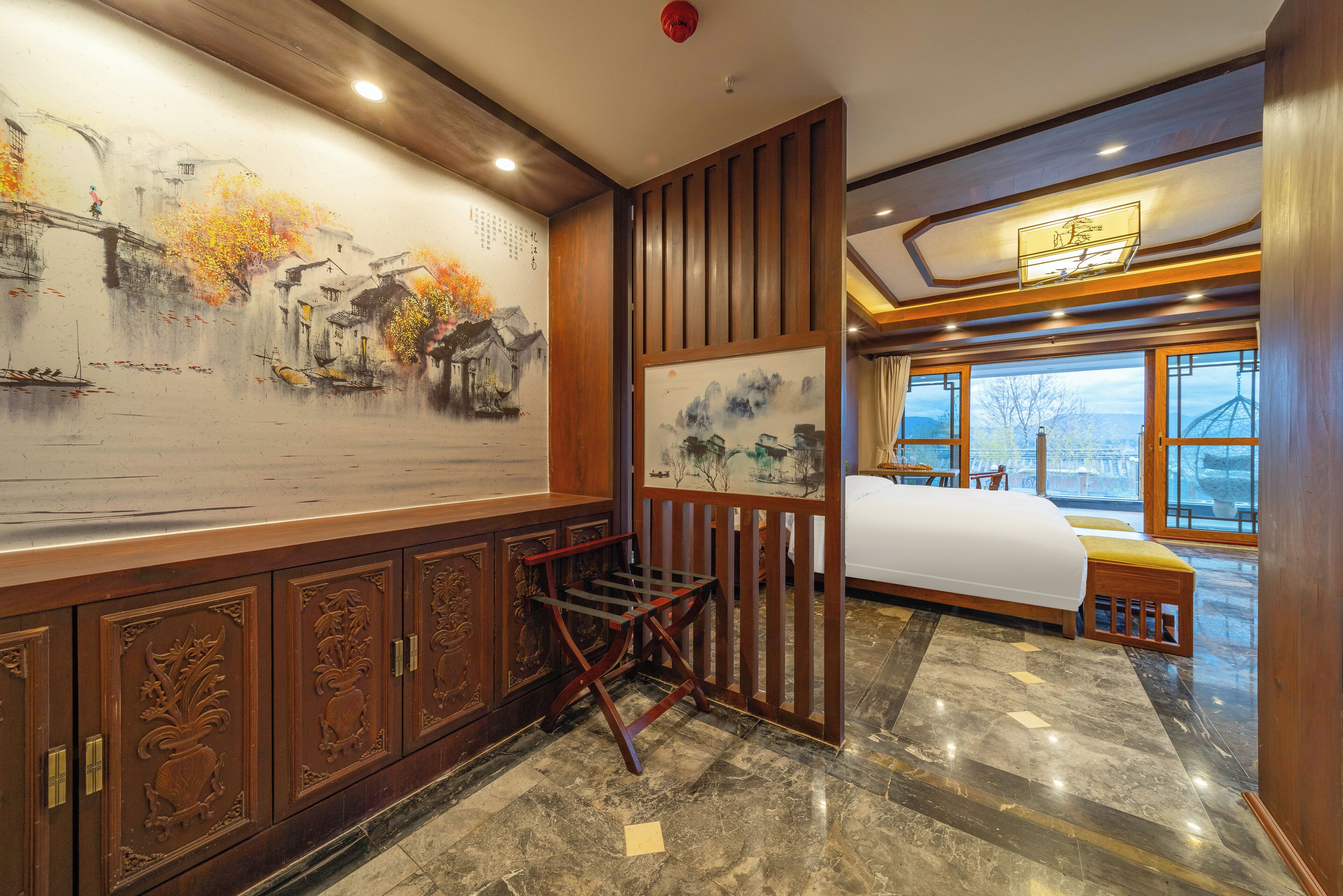 A hotel room with a large mural, a bed, a wooden bench, and a view of the outside through large windows.
