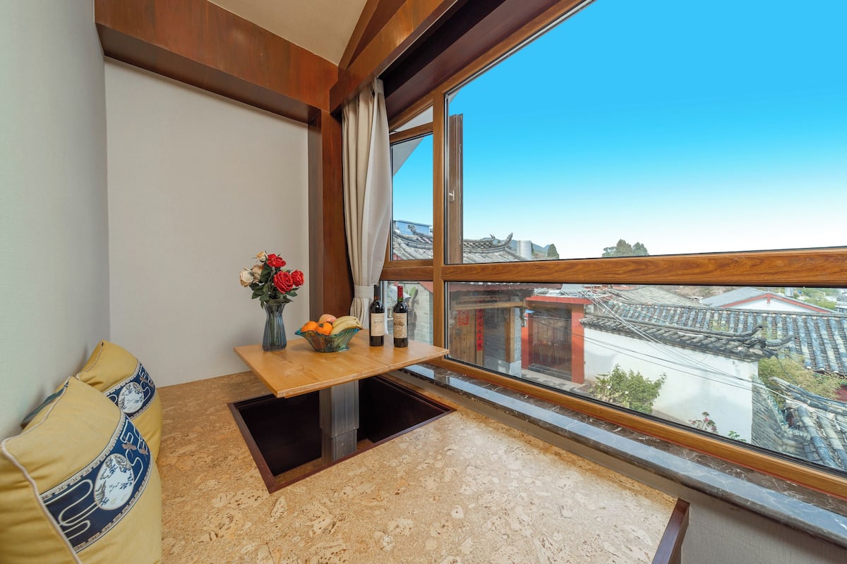 A corner room with a wooden bench, a small table with a vase of flowers, and a view of traditional buildings through a large window.