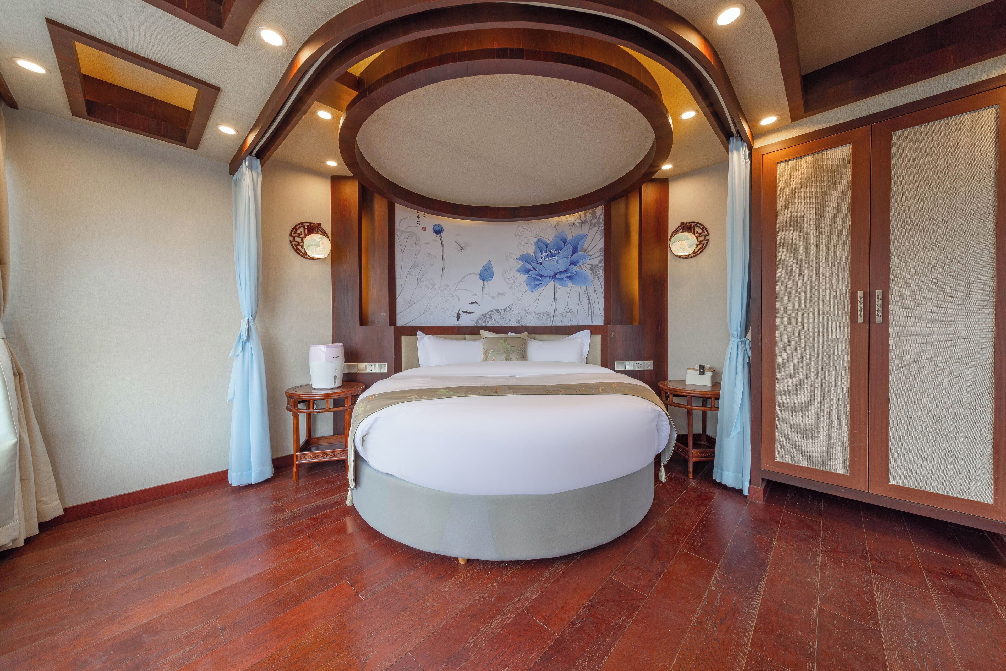 A circular bed with a headboard, wooden flooring, and a ceiling with recessed lighting.
