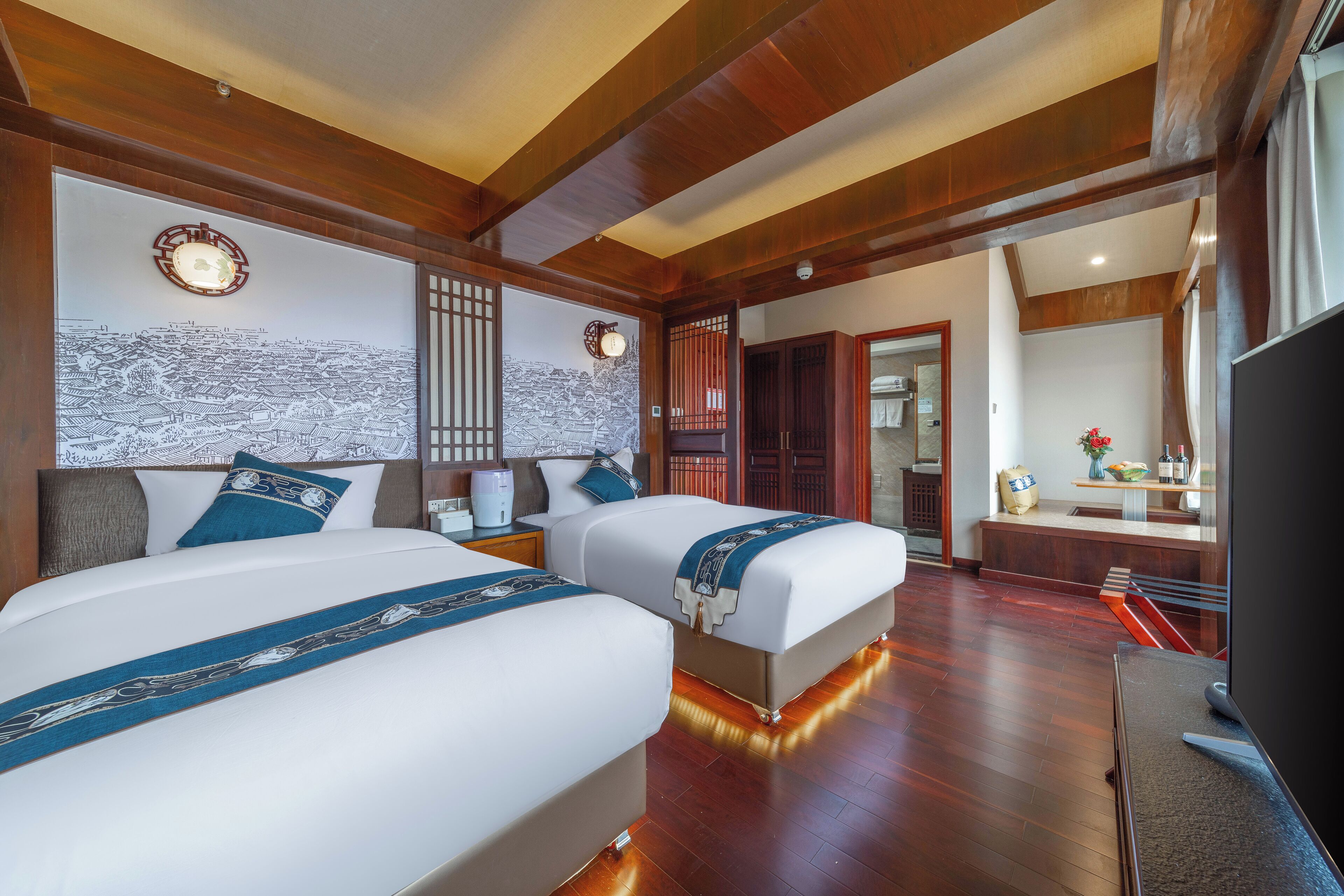 A hotel room with two beds, wooden flooring, a TV, and a bathroom visible through an open door.