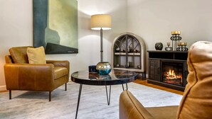 A living room with a leather sofa, a round coffee table, a floor lamp, a fireplace, and a large abstract painting. - Maine Adventure 3BR Oasis Awaits! (York)