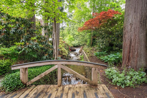 A wooden bridge over a small stream in a lush garden with various plants and trees.