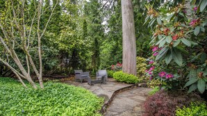 Un sendero de piedra que conduce a una zona de estar con dos sillas y una mesa, rodeada de abundante follaje y plantas con flores.