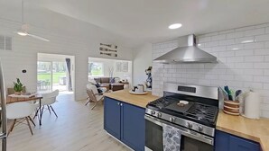 Una cocina moderna con estufa a gas, horno y salpicadero de azulejos blancos. Hay una zona de comedor con mesa y sillas, y un amplio ventanal que proporciona luz natural.