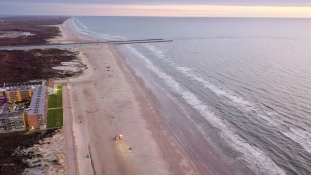 Une vue aérienne d’une plage avec une longue jetée qui s’avance dans l’eau, une plage de sable et un ensemble de bâtiments comprenant plusieurs étages et des balcons.