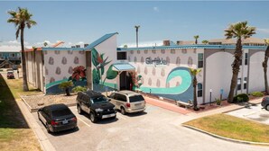 Un bâtiment orné d’une fresque représentant des feuilles et des fleurs tropicales, un parking avec trois voitures et des palmiers.