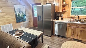 A cozy cabin interior with a wooden dining table, a sofa, a refrigerator, a dishwasher, and a window with a view of trees.