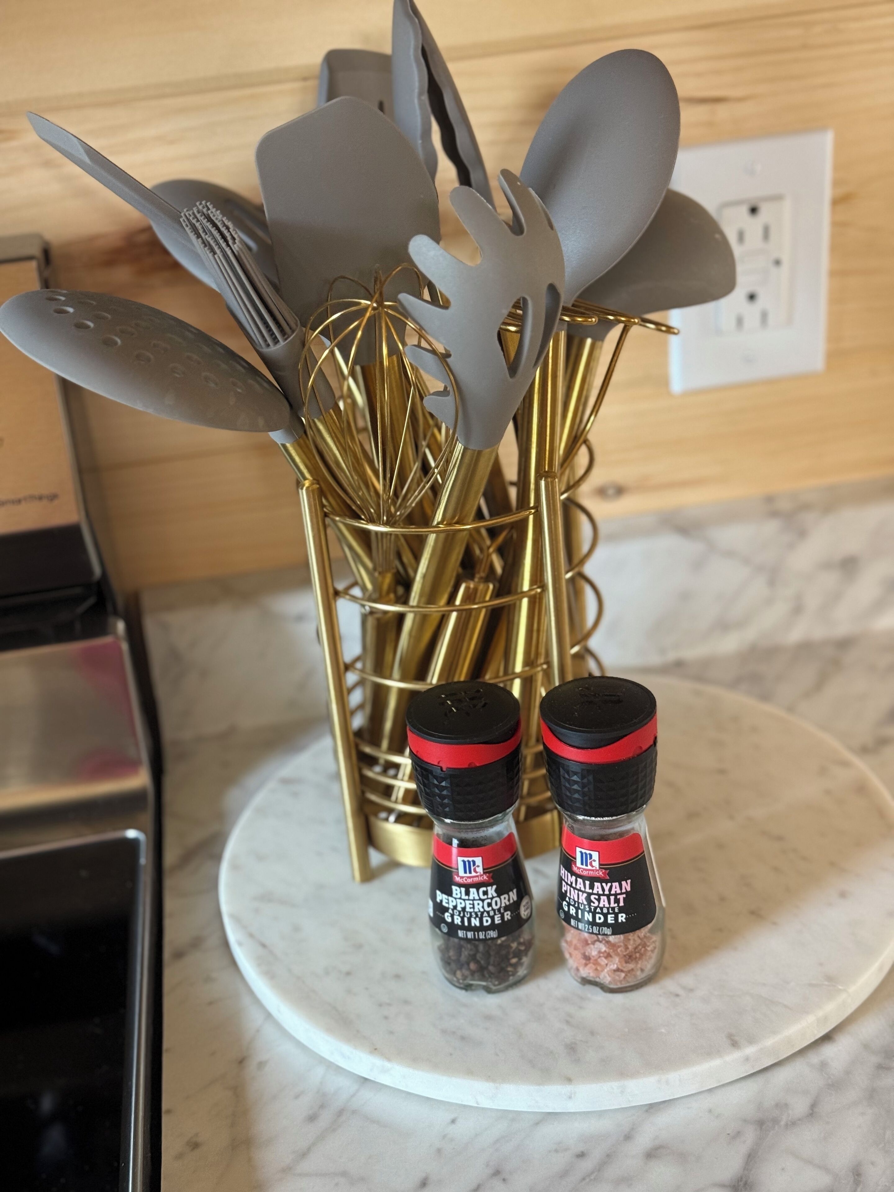 Una encimera de cocina con superficie de mármol blanco, un portalata dorada con diversos utensilios de cocina y dos frascos de especias etiquetados como McCormick Black Peppercorn Grinder y Himalayan Pink Salt Grinder.