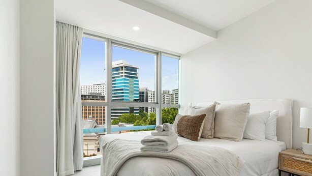 Una habitación de hotel moderna con una cama grande, una mesita de noche y vistas a un paisaje urbano a través de ventanales de piso a techo.