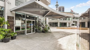 Un edificio moderno con entrada de vidrio, plantas en macetas y una torre al fondo.
