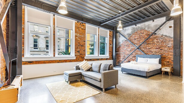 Un dormitorio tipo loft moderno con paredes de ladrillo visto, ventanas amplias, un sofá gris y una cama.