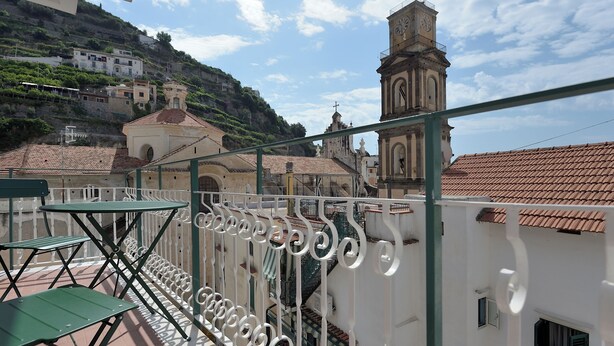 A balcony with a metal railing, a green chair, and a white building with a bell tower in the background. - The Bell Tower Clock vacation home (Minori)