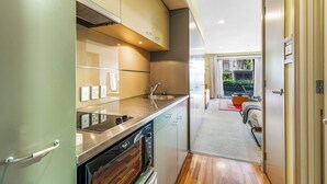 Cocina compacta con electrodomésticos modernos, un lavamanos a la vista y una puerta que da a un dormitorio con cama y una mesita.