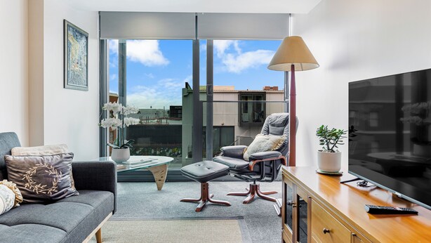 A modern living room with a sofa, armchair, ottoman, coffee table, and a TV on a wooden cabinet. - Zodiak Stays: Cozy CBD 2BR Near Top Dining Spots (Auckland)