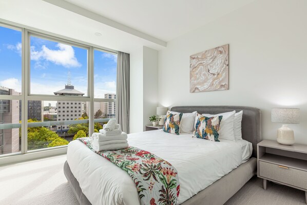 Habitación de hotel moderna con un amplio ventanal, una cama bien hecha, una mesita de noche con lámpara y vistas a la ciudad.