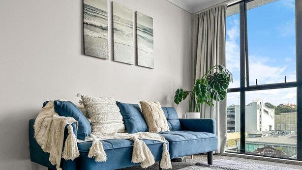 A modern living room with a blue sofa, decorative pillows, and a large window offering a view of buildings. - 2BR Newmarket Retreat near Broadway: Zodiak Stays (Auckland)