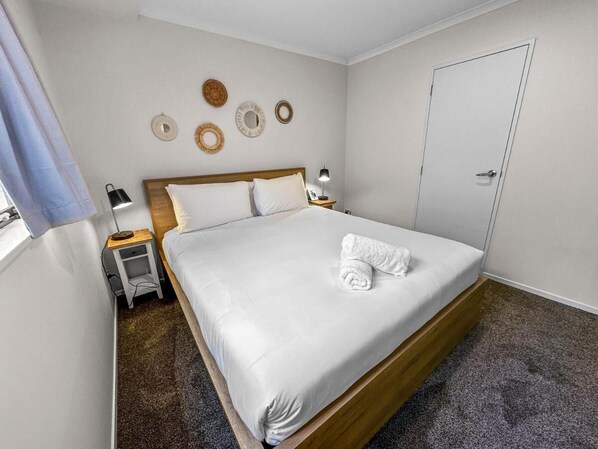 A neatly made bed with white linens and a rolled towel, a bedside table with a lamp, and a door in the room.