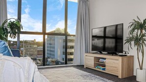 A modern bedroom with a large window, a flat-screen TV, a bed, and a plant. - 2BR Newmarket Retreat near Broadway: Zodiak Stays (Auckland)