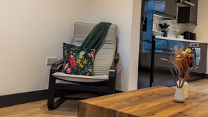 Ein modernes Wohnzimmer mit einem grauen Sofa, einem Holztisch und einer Vase mit getrockneten Blumen. Im Hintergrund ist eine Küche mit Edelstahlgeräten erkennbar.