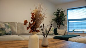 Ein Holztisch mit zwei weißen Vasen, gefüllt mit getrockneten Pflanzen und Schilfhalmen. Im Hintergrund steht ein beiges Sofa mit grünen Kissen.