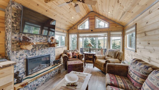 A cozy cabin interior with a stone fireplace, wooden walls, a ceiling fan, and large windows offering views of the outdoors. - Mountain Views: Cabin in Fort Garland Near Hiking (Fort Garland)