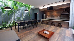A modern kitchen with a wooden countertop and bar stools, a dining area with a wooden table and chairs, and a large window with tropical plants.