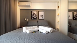Ein Hotelzimmer mit einem großen Bett, grauer Bettwäsche, zwei Kissen, einem Nachttisch, einer Wand-Klimaanlage und zwei gerahmten Kunstwerken an der Wand.