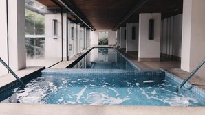 A modern indoor swimming pool with clear glass walls, a wooden ceiling, and a view of greenery outside.