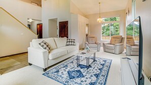 Ein geräumiges Wohnzimmer mit einem weißen Sofa, zwei Sesseln, einem Couchtisch und einem großen Flachbildfernseher. Im Hintergrund ist eine Treppe mit Deckenventilator und einer Holztür zu sehen.