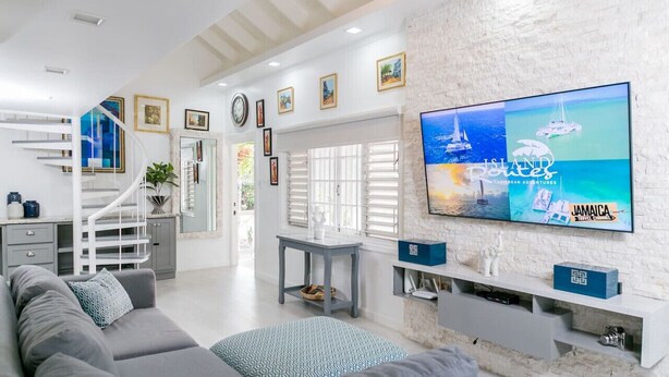 A modern living room with a TV displaying 'Island Paradise Jamaica Travel Channel', a grey sofa, a white staircase, and a white brick wall. - Choose To Be Happy @ The Seville #E3 [2-Bedroom] (Kingston)