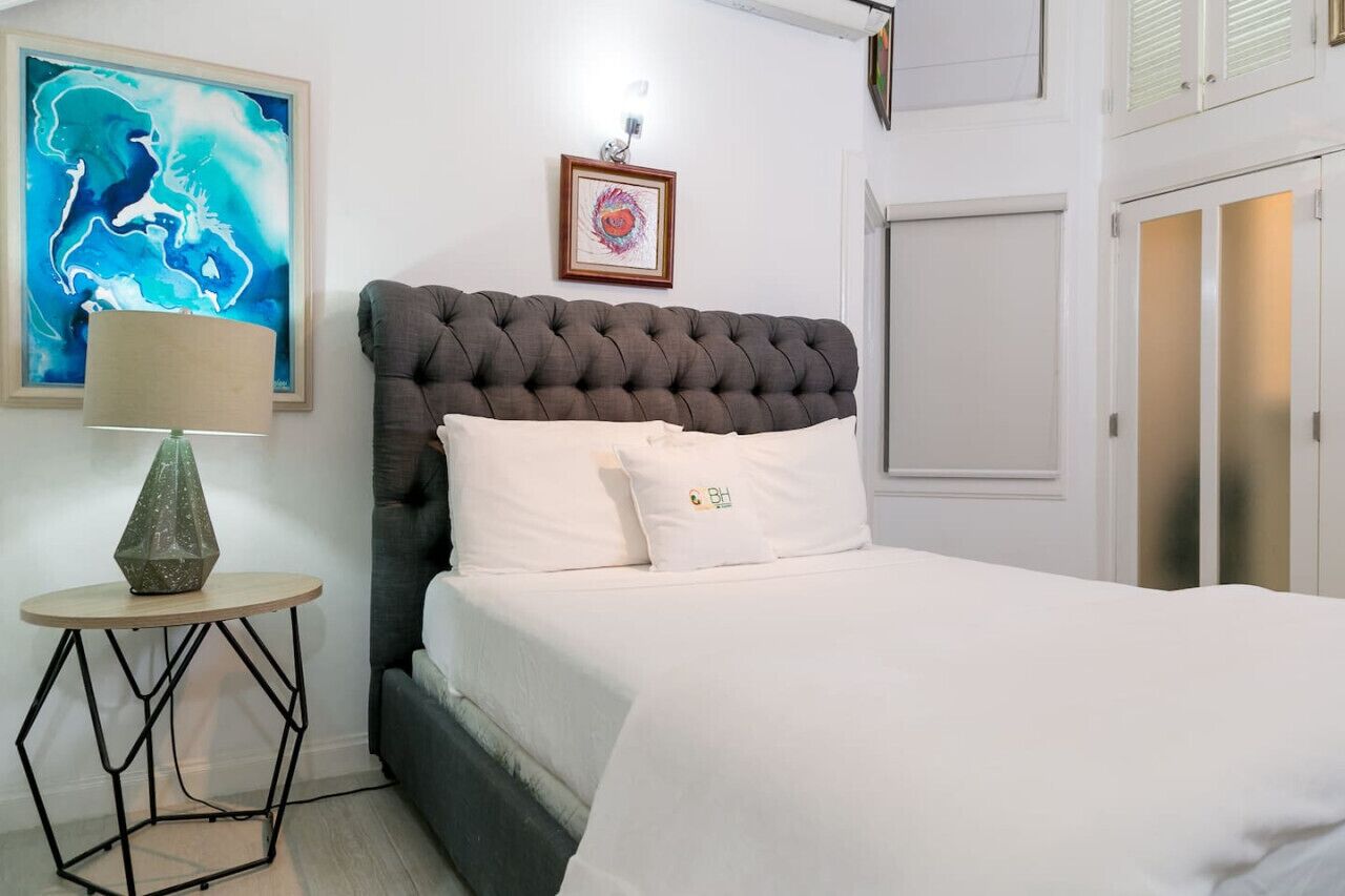 Ein ordentlich eingerichtetes Schlafzimmer mit einer gepolsterten Kopfteil, einem Bett mit weißer Bettwäsche, einem runden Nachttisch mit Lampe und einem gerahmten Bild an der Wand.