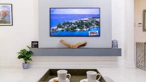A modern hotel lobby with a large TV displaying a scenic view of Jamaica, a wooden decorative piece, a tray with cups and cookies, and a sign about smoking and vaping. - Choose To Be Happy @ The GAIA [2-Bedroom w/ Pool] (Kingston)