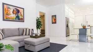 A modern living room with a sectional sofa, a small ottoman, and a coffee table. There is a potted plant and artwork on the walls. - Choose To Be Happy @ The GAIA [2-Bedroom w/ Pool] (Kingston)