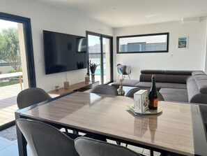Ein modernes Wohnzimmer mit einem großen Flachbildfernseher, einem Esstisch, grauen Sofas und Blick ins Freie.