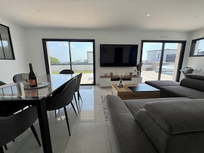 A modern living room with a dining area, a flat-screen TV, and large windows offering a view of the outdoors.