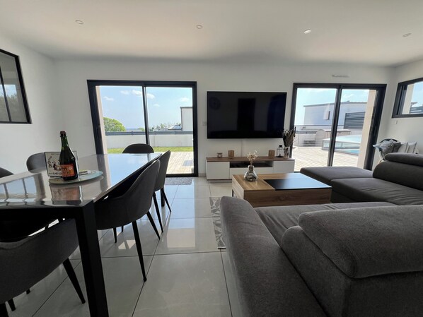 Ein modernes Wohnzimmer mit Essbereich, einem Flachbildfernseher und großen Fenstern mit Blick ins Freie.
