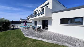 Een modern huis met twee verdiepingen, een houten terras, zitplaatsen buiten en een zwembad.