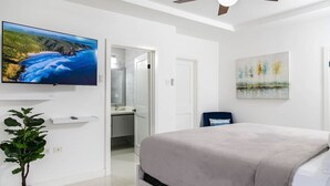 A bedroom with a bed, a TV mounted on the wall, a plant, and a painting.