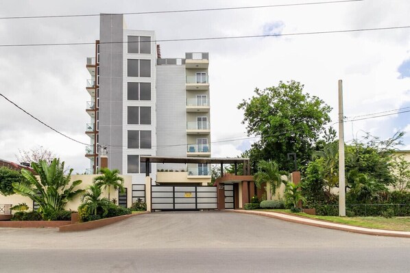A modern multi-story residential building with a gated entrance, surrounded by greenery and a clear sky.