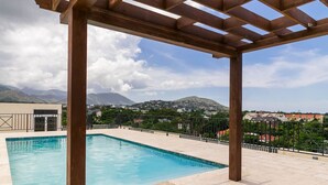 A rooftop pool with a pergola structure, surrounded by a fence, overlooking a mountainous landscape.