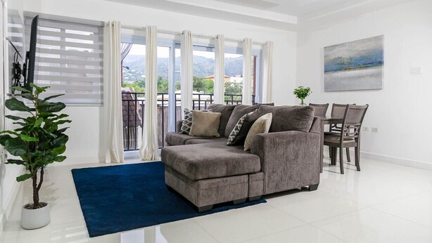 A modern living room with a grey sofa, a dining table with chairs, a ceiling fan, and a large window with a view of mountains.