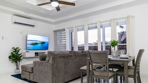 A modern living room with a grey sofa, dining table, and chairs. There is a flat-screen TV mounted on the wall, a ceiling fan, and a potted plant.
