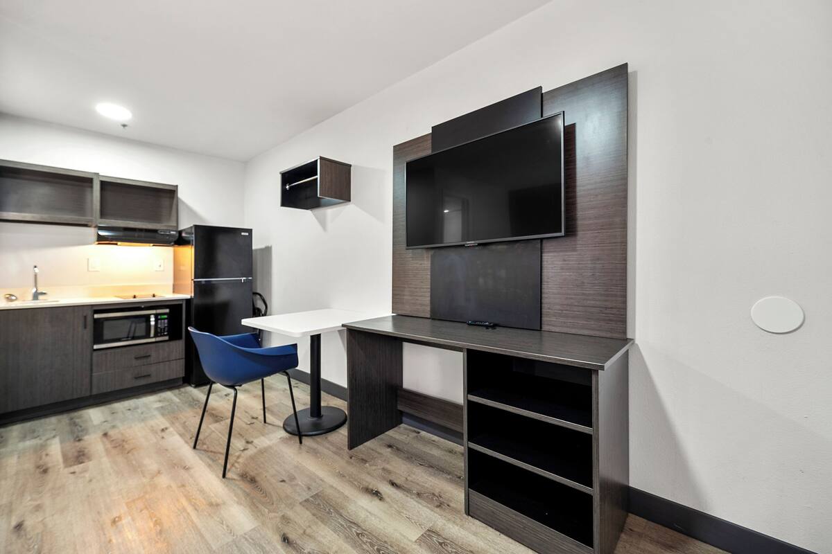 A modern kitchen with a built-in microwave, oven, and refrigerator. There is a dining table with a blue chair and a flat-screen TV mounted on the wall.