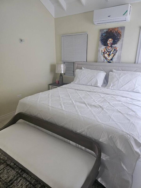 A neatly made bed with a white quilt, a bedside table with a lamp, and a wall-mounted air conditioner.